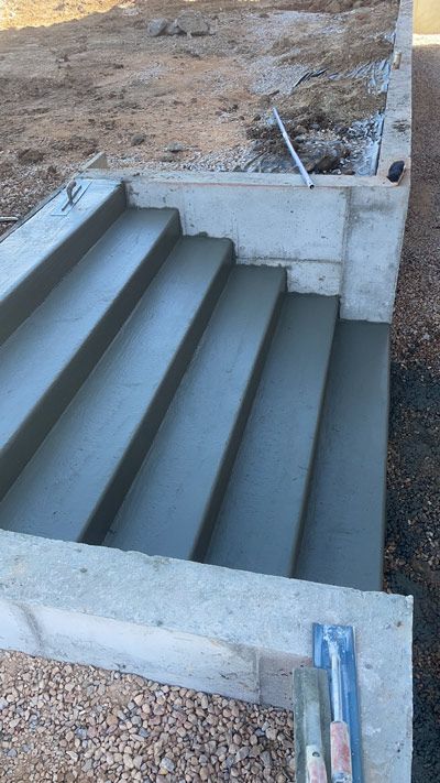 Concrete steps with dark treads at a construction site, bordered by unfinished walls and dirt.