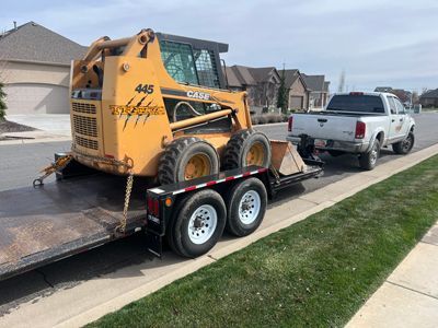 Yellow skid steer on a trailer being towed by a white pickup truck on a suburban street.