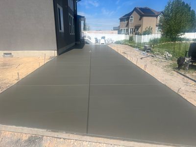 Concrete walkway beside a dark house in a suburban neighborhood under construction, leading toward homes and trees.