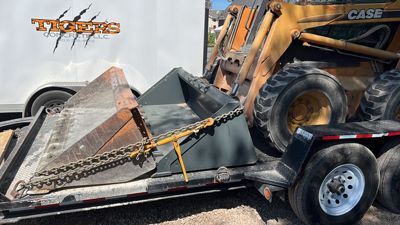 Front loader lifting a crushed black trailer with chains, beside a white truck and CASE machine