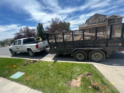 White pickup truck towing a flatbed trailer parked on a residential street beside a sidewalk and lawn.