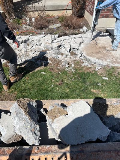 Workers removing broken concrete around a lawn beside a house, with rubble scattered on the ground