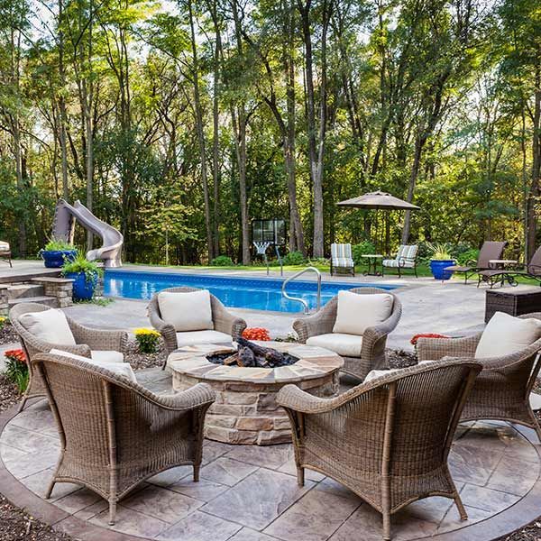 Backyard pool with wicker chairs around a stone fire pit, surrounded by trees and patio umbrellas