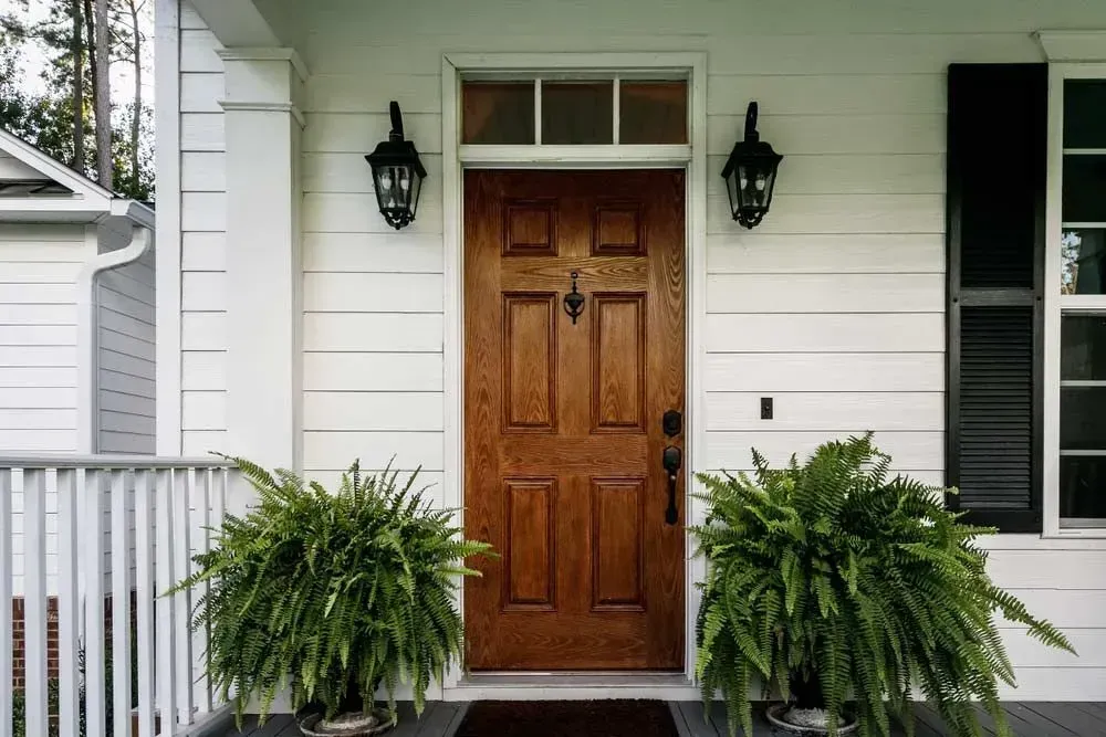 Wooden front door with ferns, black sconces, and white siding.