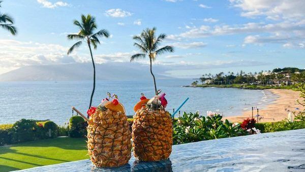 Two pineapple cocktails on a ledge, overlooking a beach and ocean. Palm trees, blue sky.