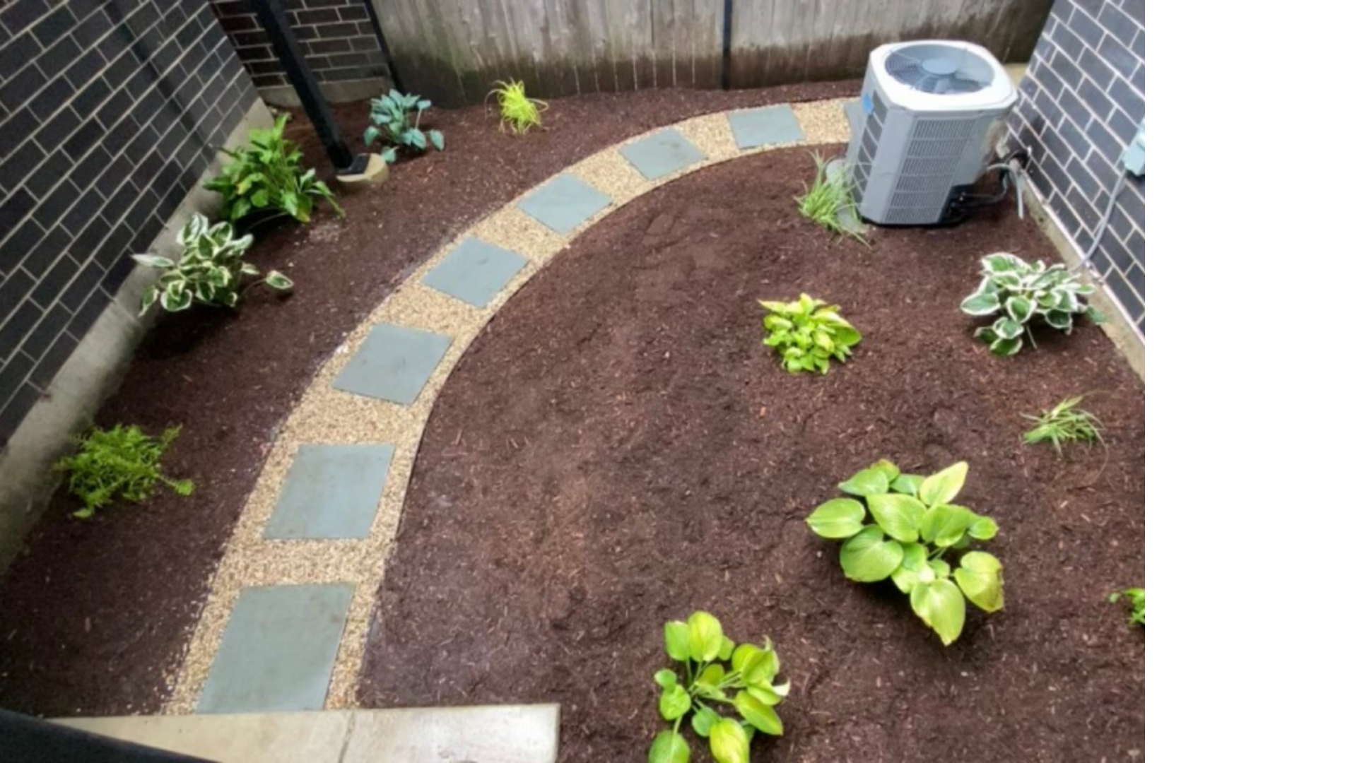 A small backyard with a stone path, mulch, and plants. An air conditioning unit sits in the corner.