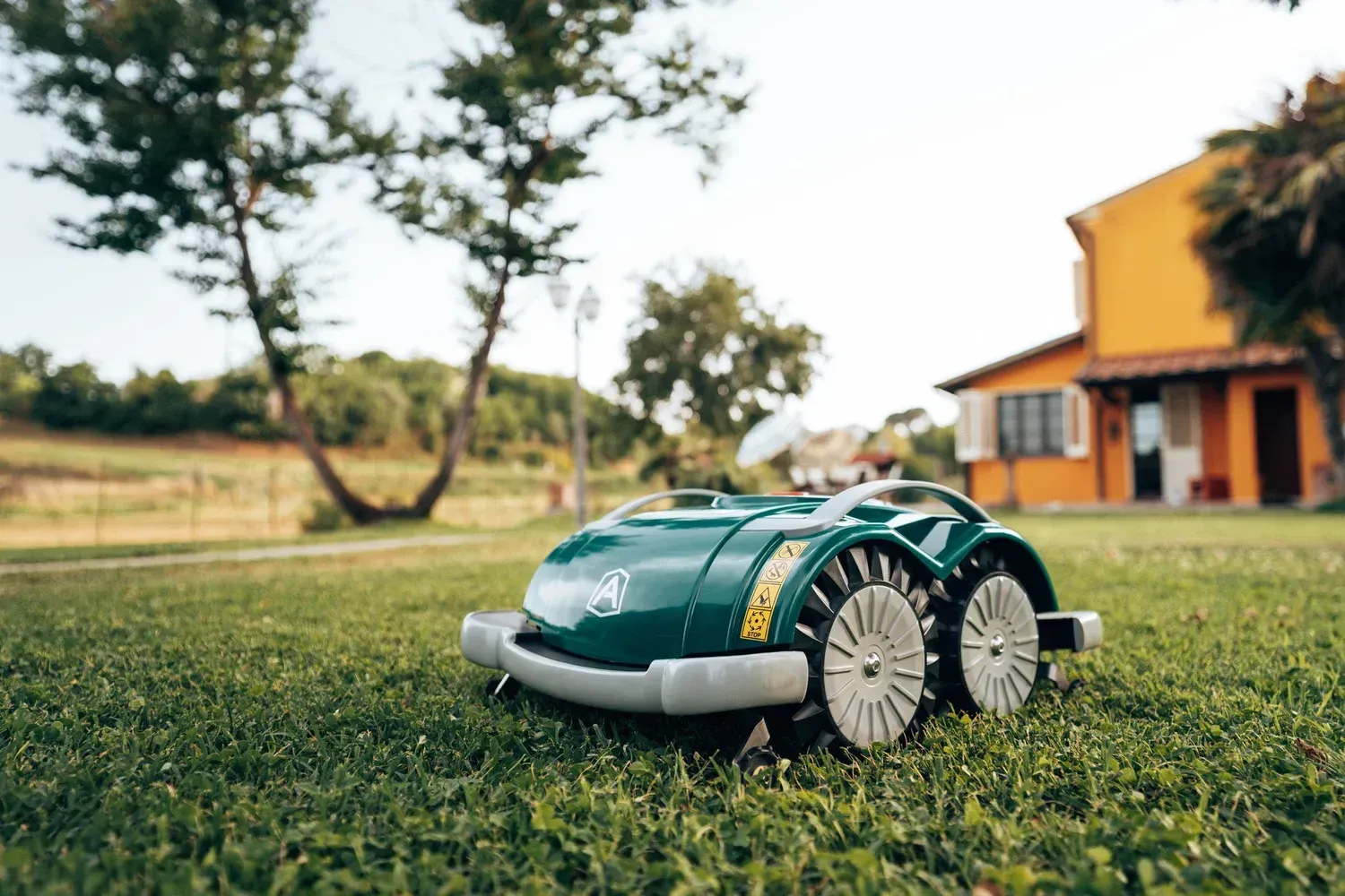 Green robotic lawnmower on a grassy lawn with a house in the background.