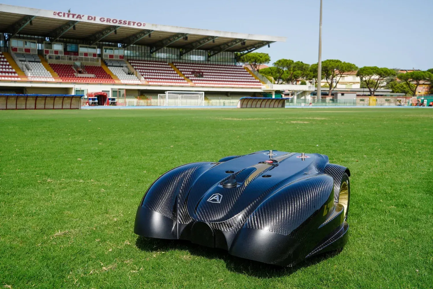 Black robotic lawnmower on a green field in front of a stadium with red and white seating.