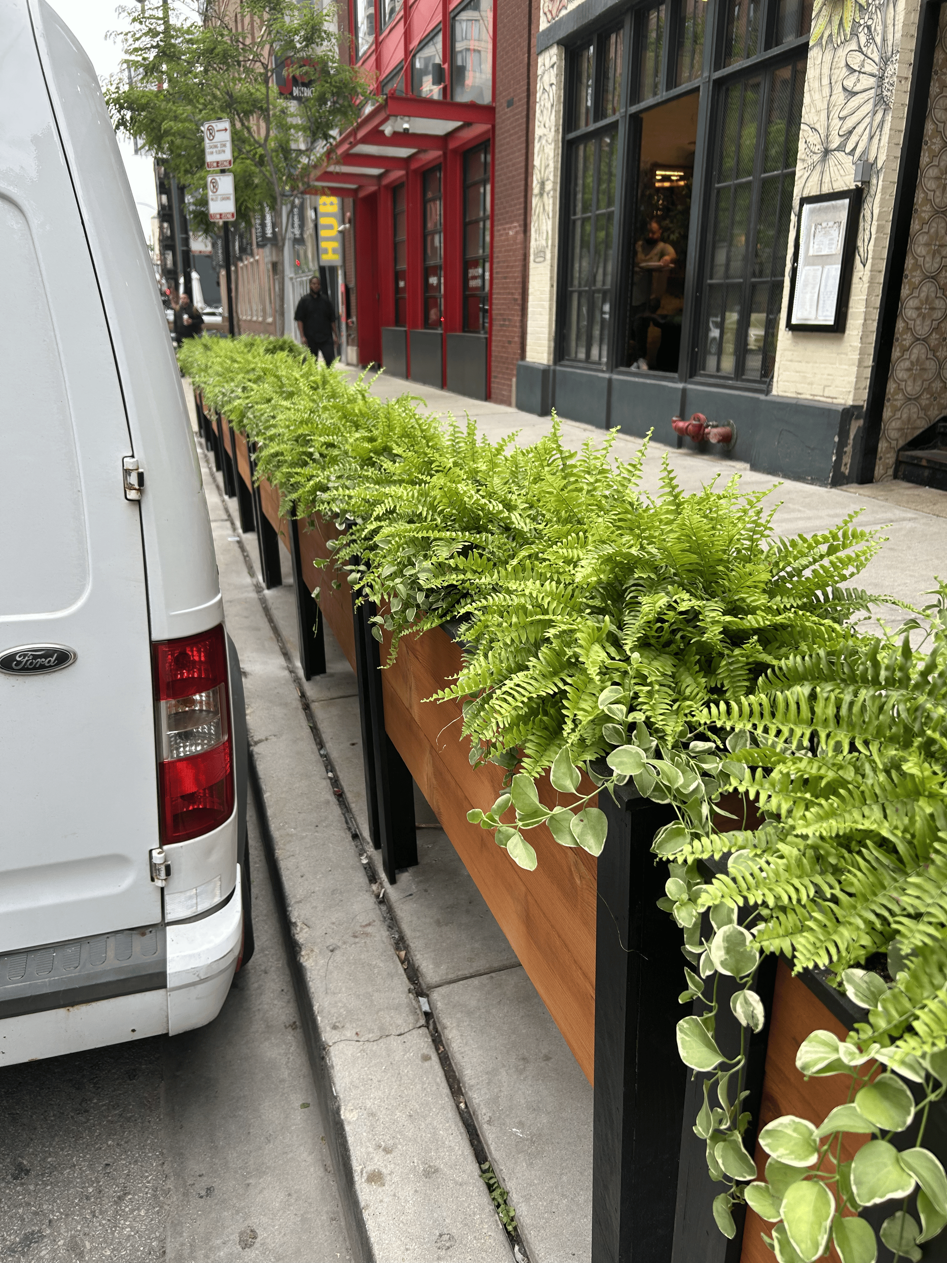 White van parked next to a long wooden planter filled with green plants on a sidewalk by buildings.
