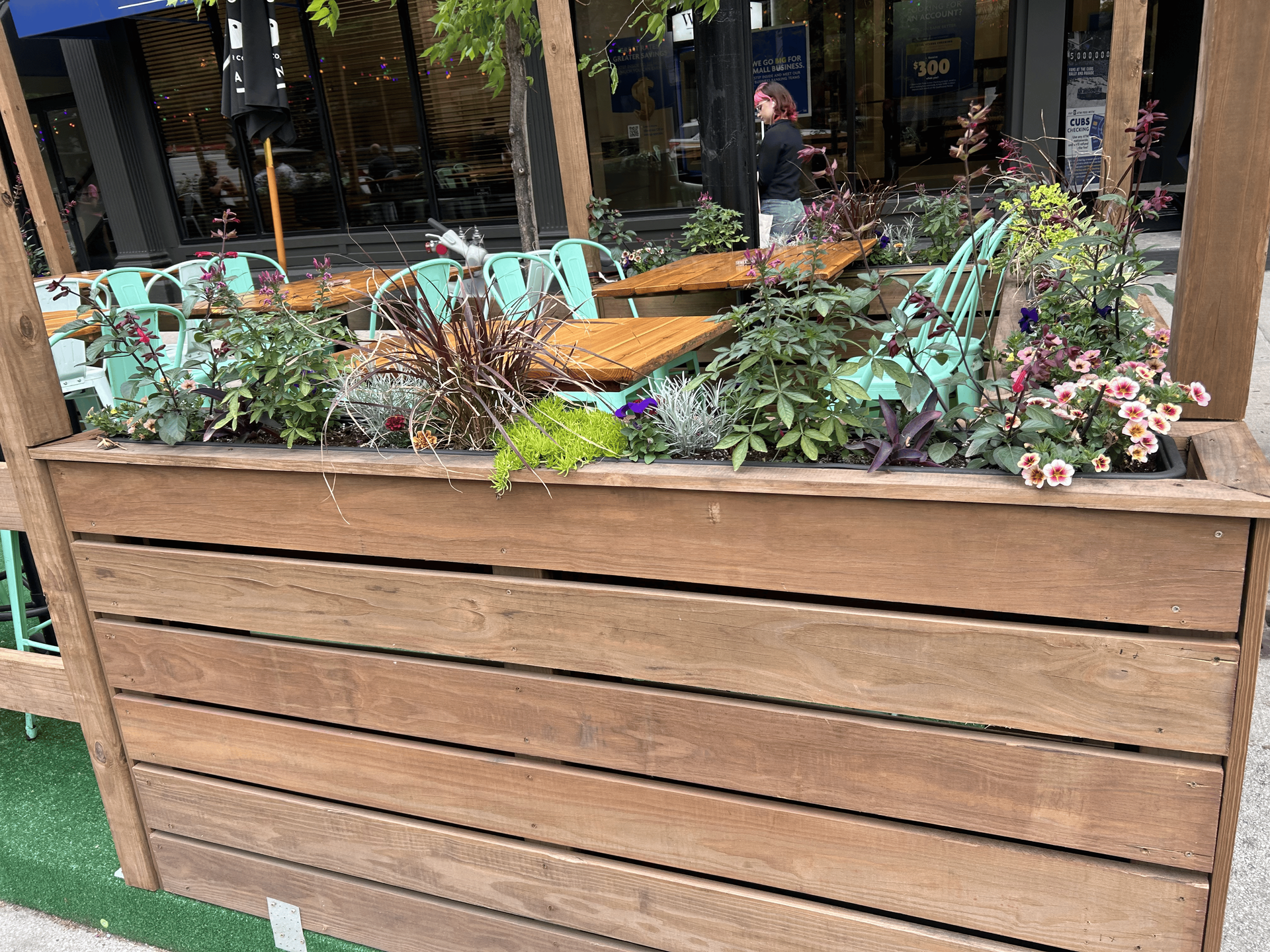 Wooden planter box with various plants in front of an outdoor seating area.