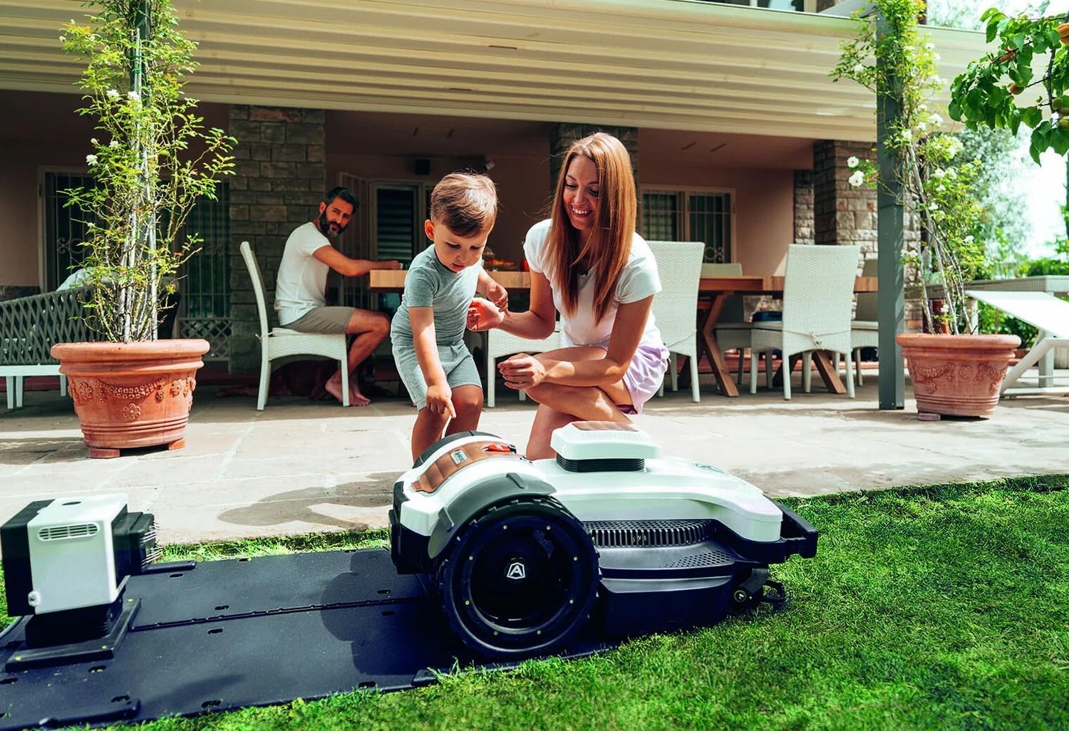 Woman and child with a robotic lawnmower, man in background. Exterior setting with grass and patio.