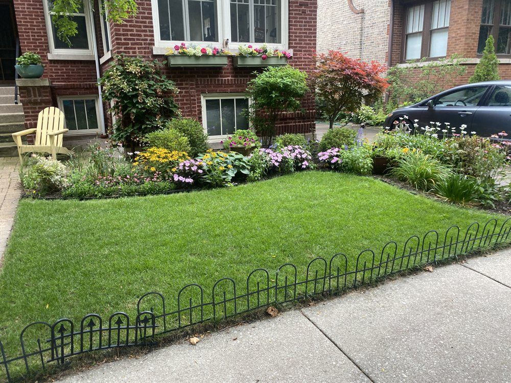 Lush front yard with manicured lawn, surrounded by a black fence and colorful flowerbeds. Brick building in the background.