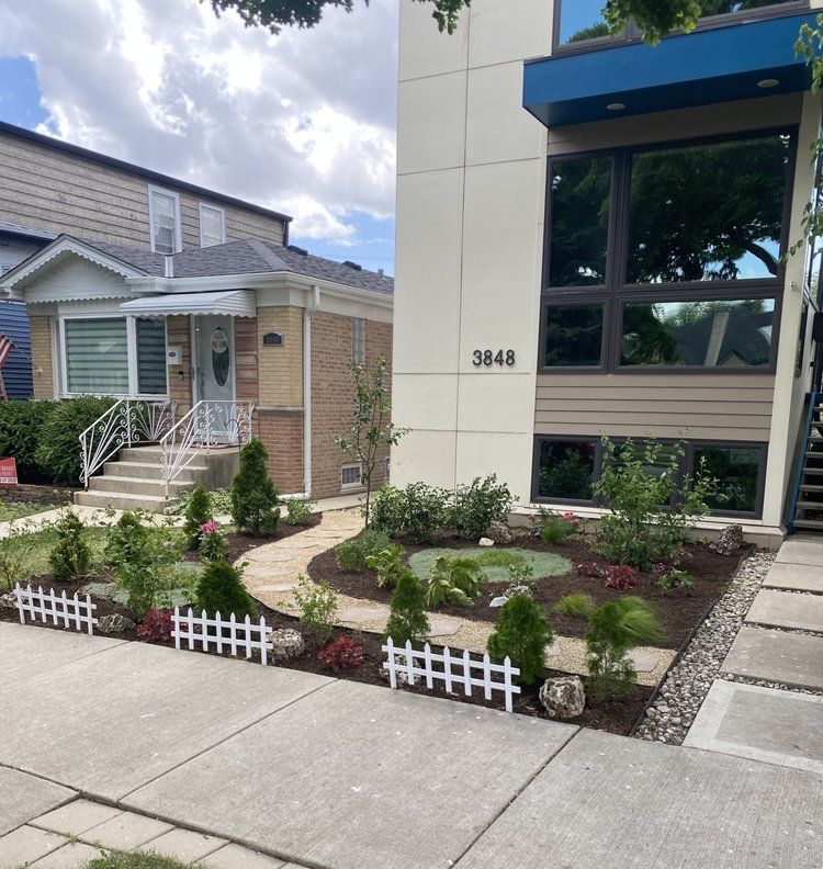 Street view of a modern house (3848) with a small garden. A brick house sits adjacent. A white picket fence frames the garden.