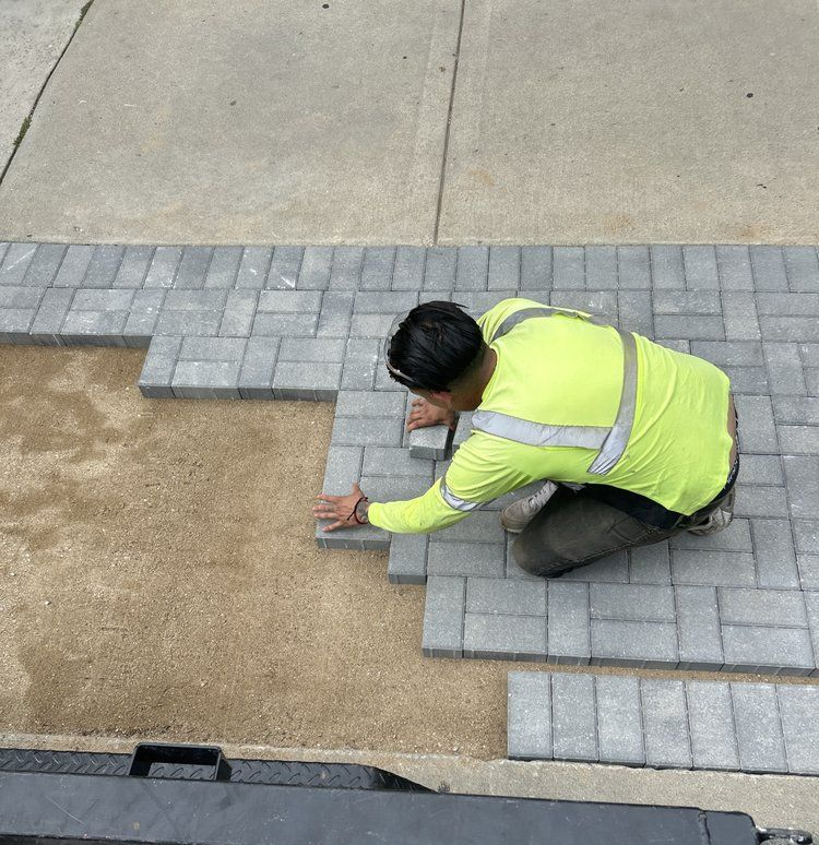 Worker installing gray paving stones on a sandy base, wearing a yellow safety vest; outdoors.