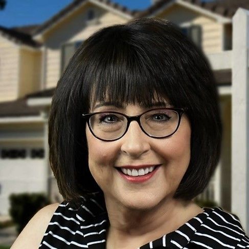 Smiling woman with dark hair and glasses, in front of a house; wearing a striped top.