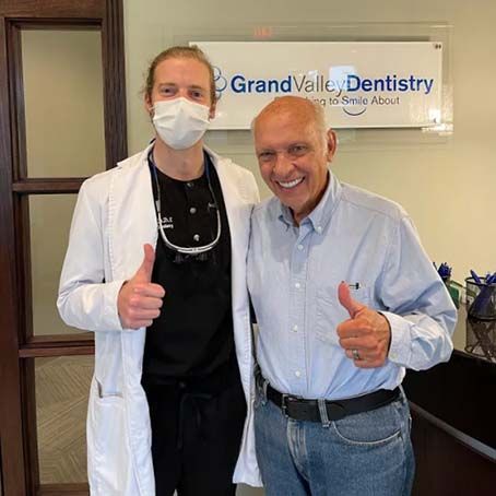 Two men standing in a dental office, smiling and giving thumbs-up, one in a white coat and mask.