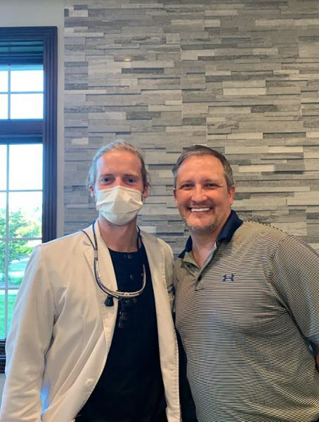 Two smiling men standing indoors in front of a gray stone wall, one wearing a mask and white coat.