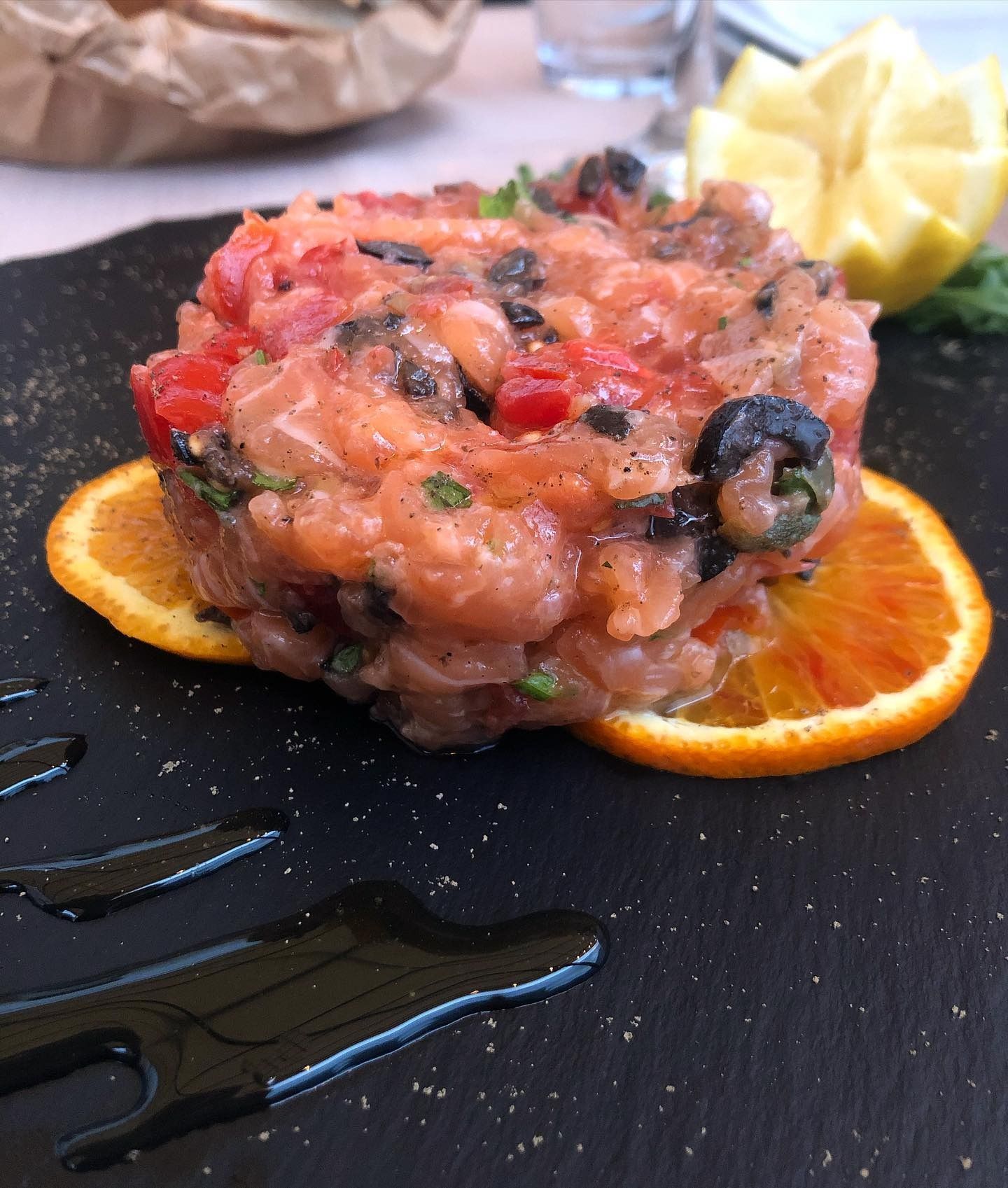 Tartare di salmone con olive nere, fette d'arancia e spicchi di limone su una lavagna nera.