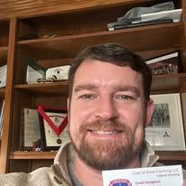 Smiling man in an office, holding up a business card in front of a bookshelf.