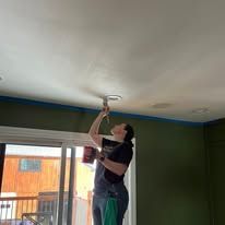 Person painting the ceiling near a window in a room with green walls and blue painter’s tape.