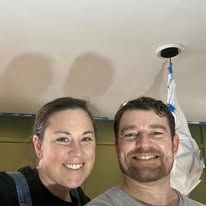 Two smiling people taking a selfie indoors, with a ceiling fixture and a hanging bag behind them.