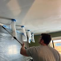 Person using a paint roller on a ceiling, with plastic-covered walls and windows in a room under renovation