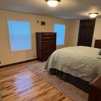 Bedroom with hardwood floor, large bed, dresser, and two windows with closed blinds.