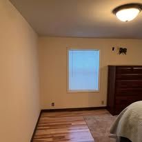 Empty beige bedroom with window, ceiling light, hardwood floor, and a bed edge on the right