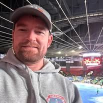 Selfie of a man in a gray hoodie at an indoor sports arena with bright lights and a crowd in the background