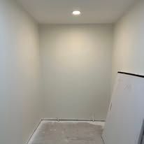 Small empty white room with a recessed ceiling light and a gray floor panel leaning against the wall.
