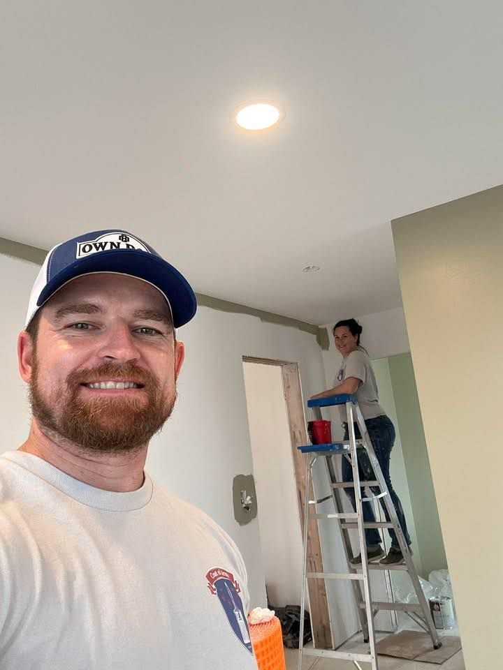 Man smiling in foreground; another person stands on a ladder painting a wall in a bright room.