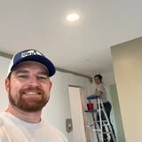 Man smiling in a blue cap poses in the foreground while another stands on a ladder painting a wall.