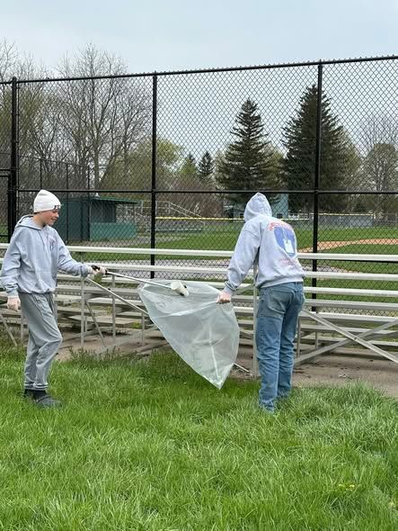 Two workers in white hoodies collect trash beside bleachers on a grassy field near a fenced sports court
