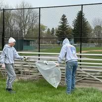 Two workers in gray hoodies cover bleachers with plastic at a fenced outdoor sports field.