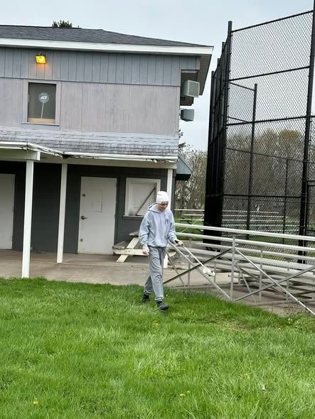 Person walking on grass beside a building and chain-link fence, carrying metal bleachers.