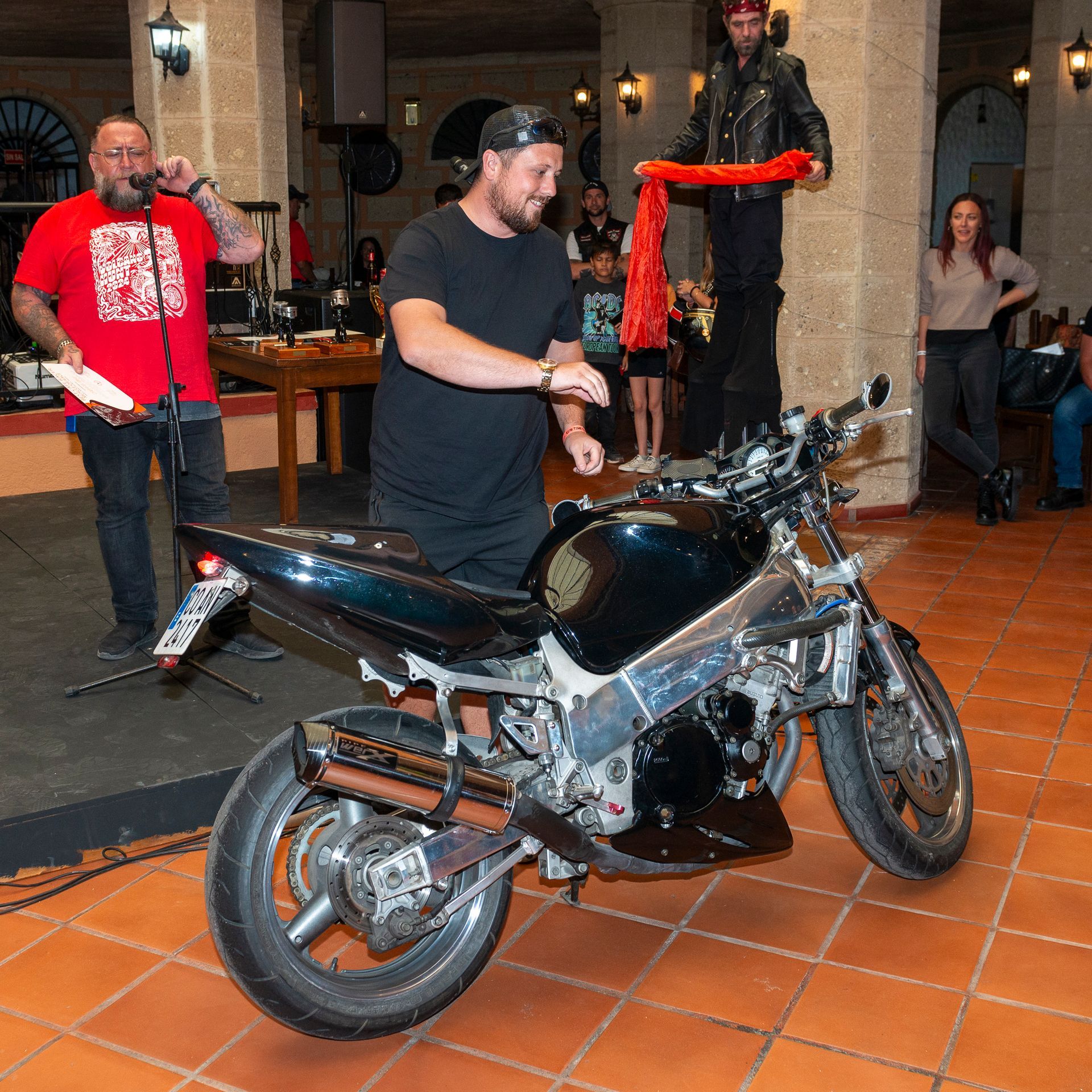 A man in a red shirt is singing into a microphone next to a motorcycle