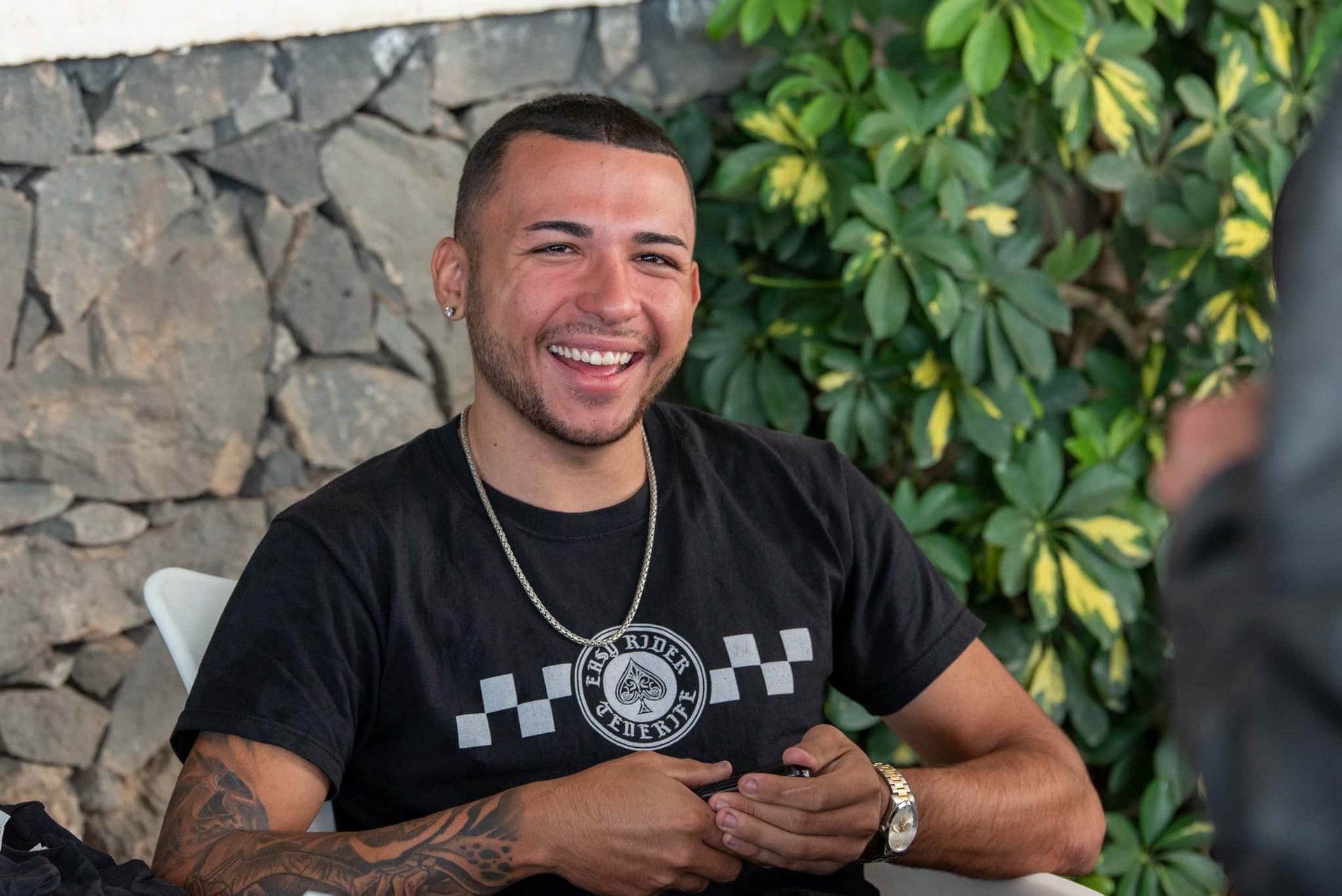 A man wearing a black shirt and a chain is smiling while sitting at a table.