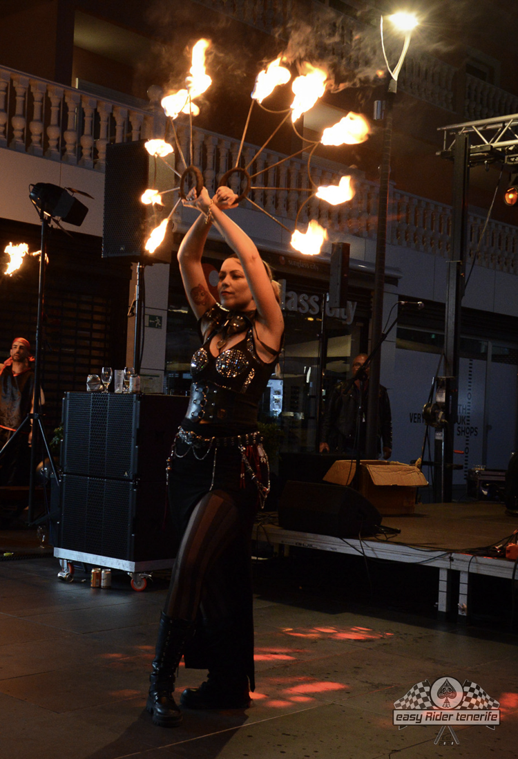 Woman in black outfit performing fire dance, holding flaming props in an outdoor setting.