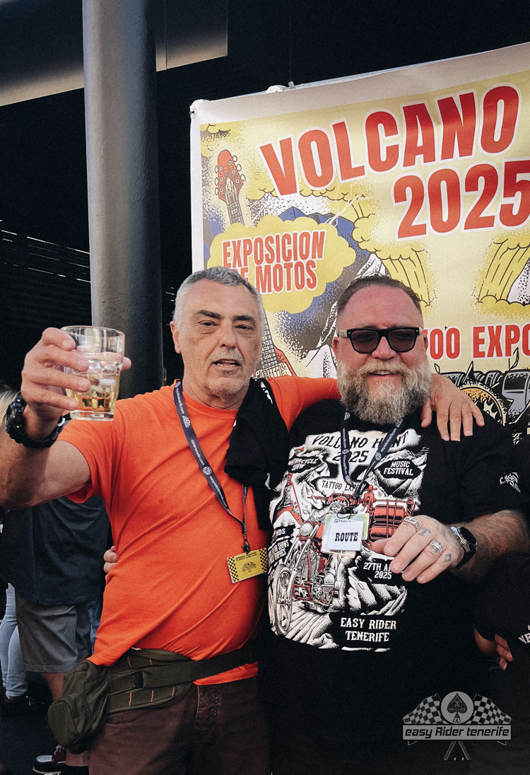 Two men are posing for a picture in front of a sign that says volcano 2025