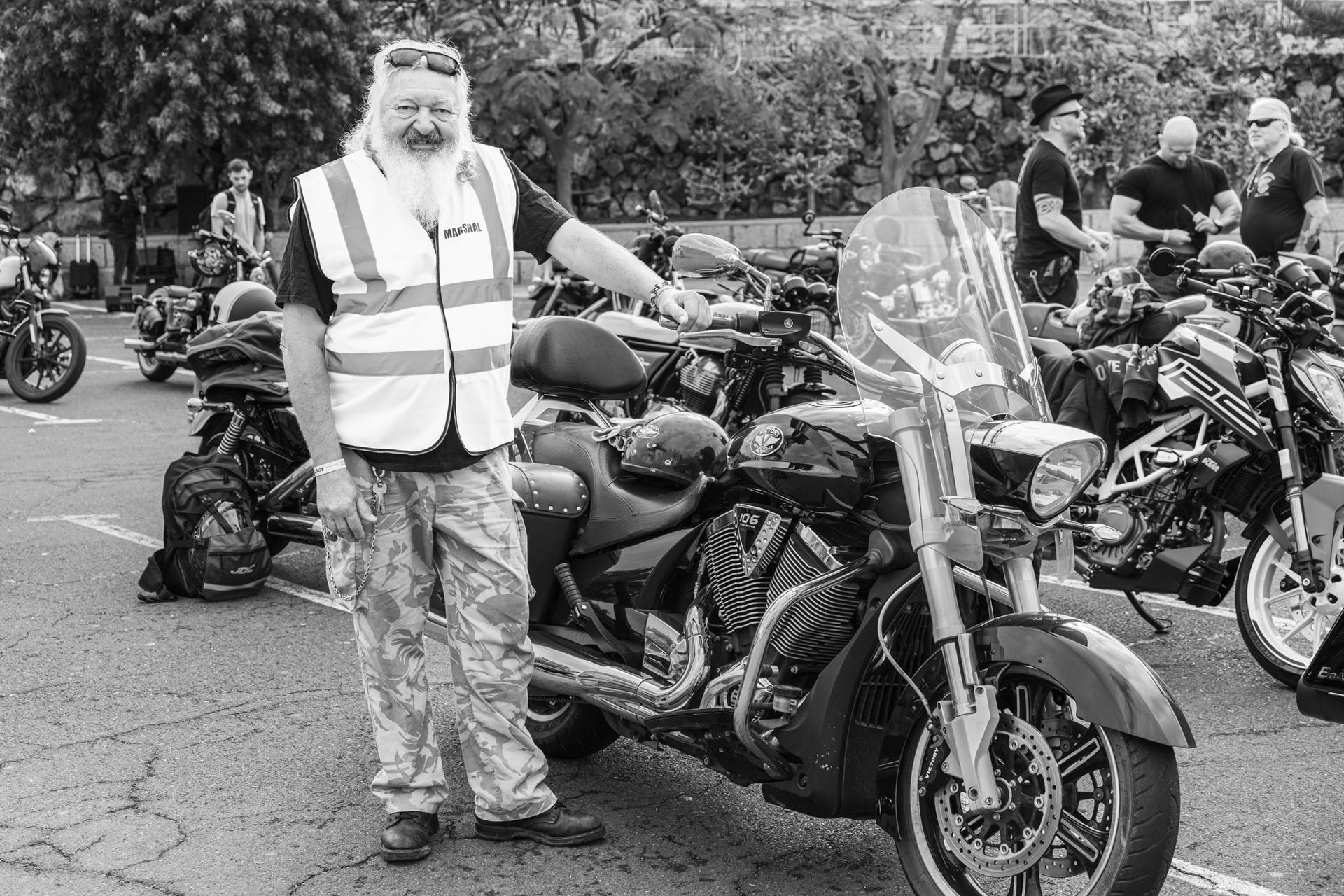 A man with a beard is standing next to a motorcycle in a parking lot.