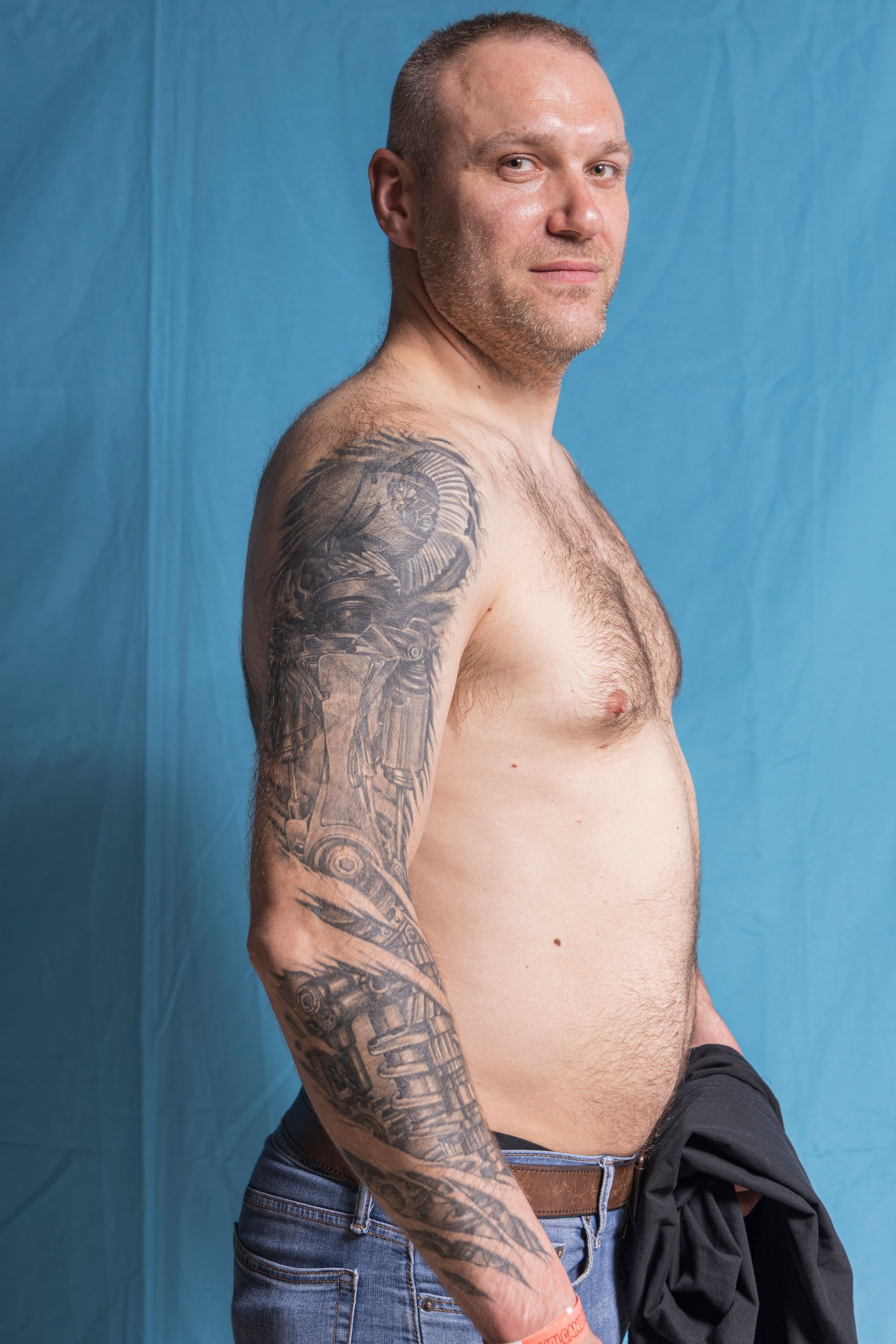 A shirtless man with tattoos on his arms is standing in front of a blue wall.