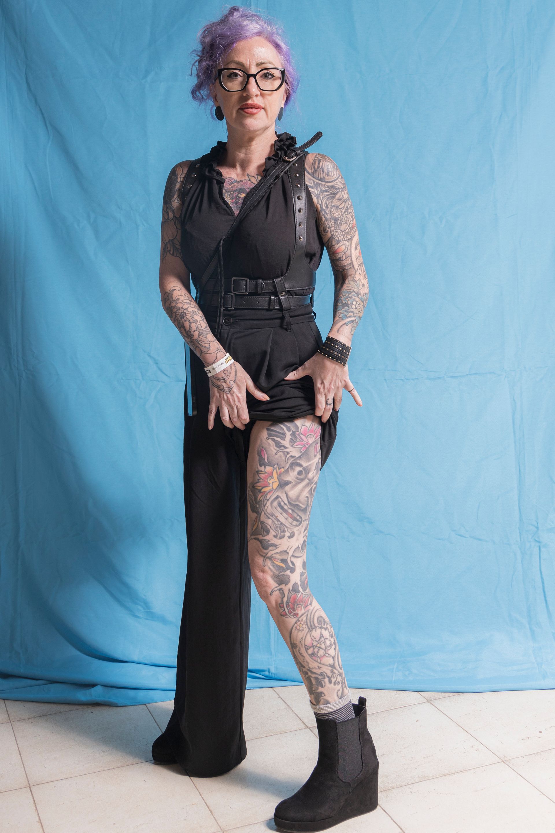 A woman with purple hair and tattoos is standing in front of a blue curtain.
