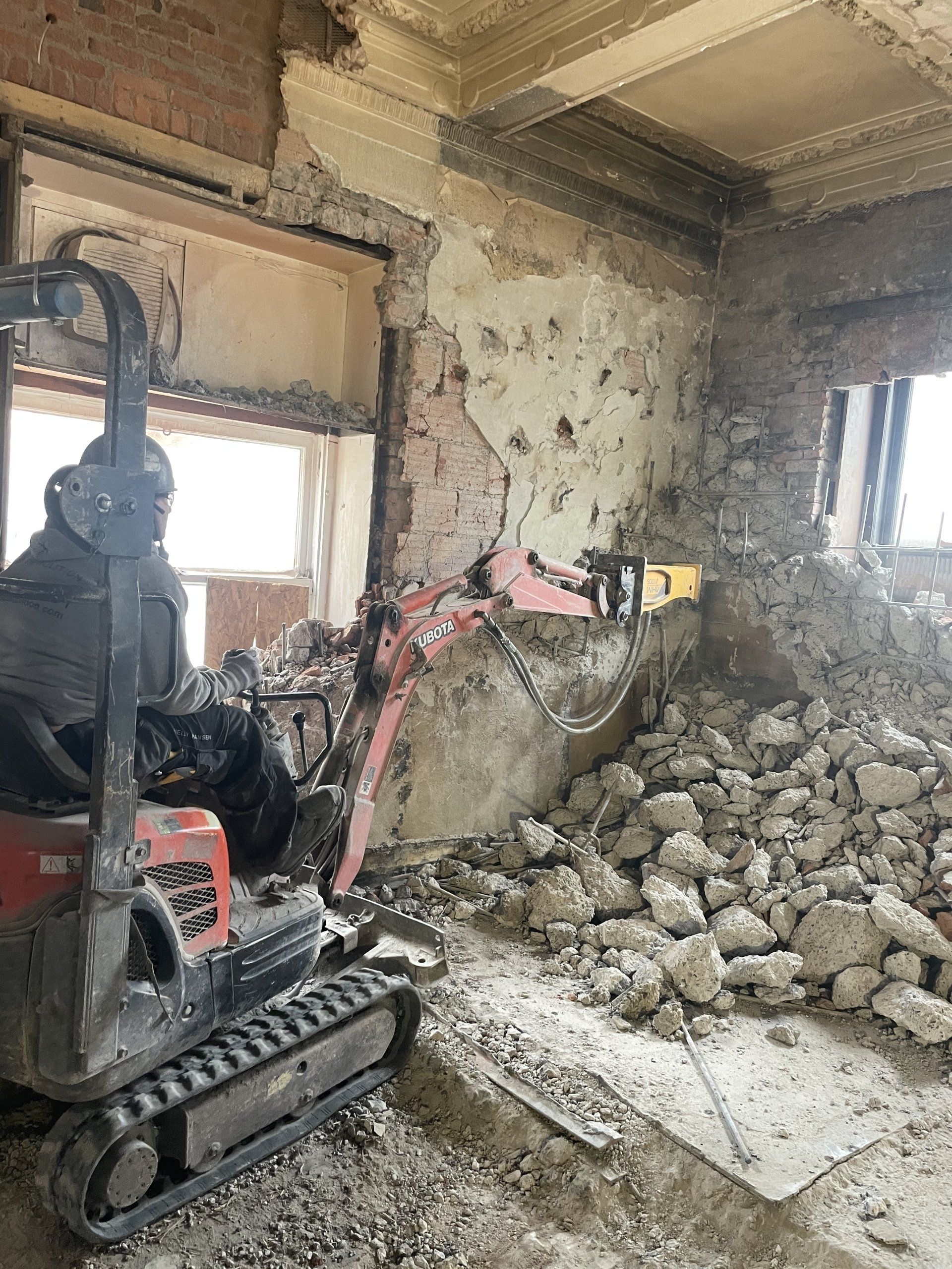 Une mini-pelle démolit un mur à l'intérieur d'un bâtiment ; les débris s'amoncellent sur le sol.