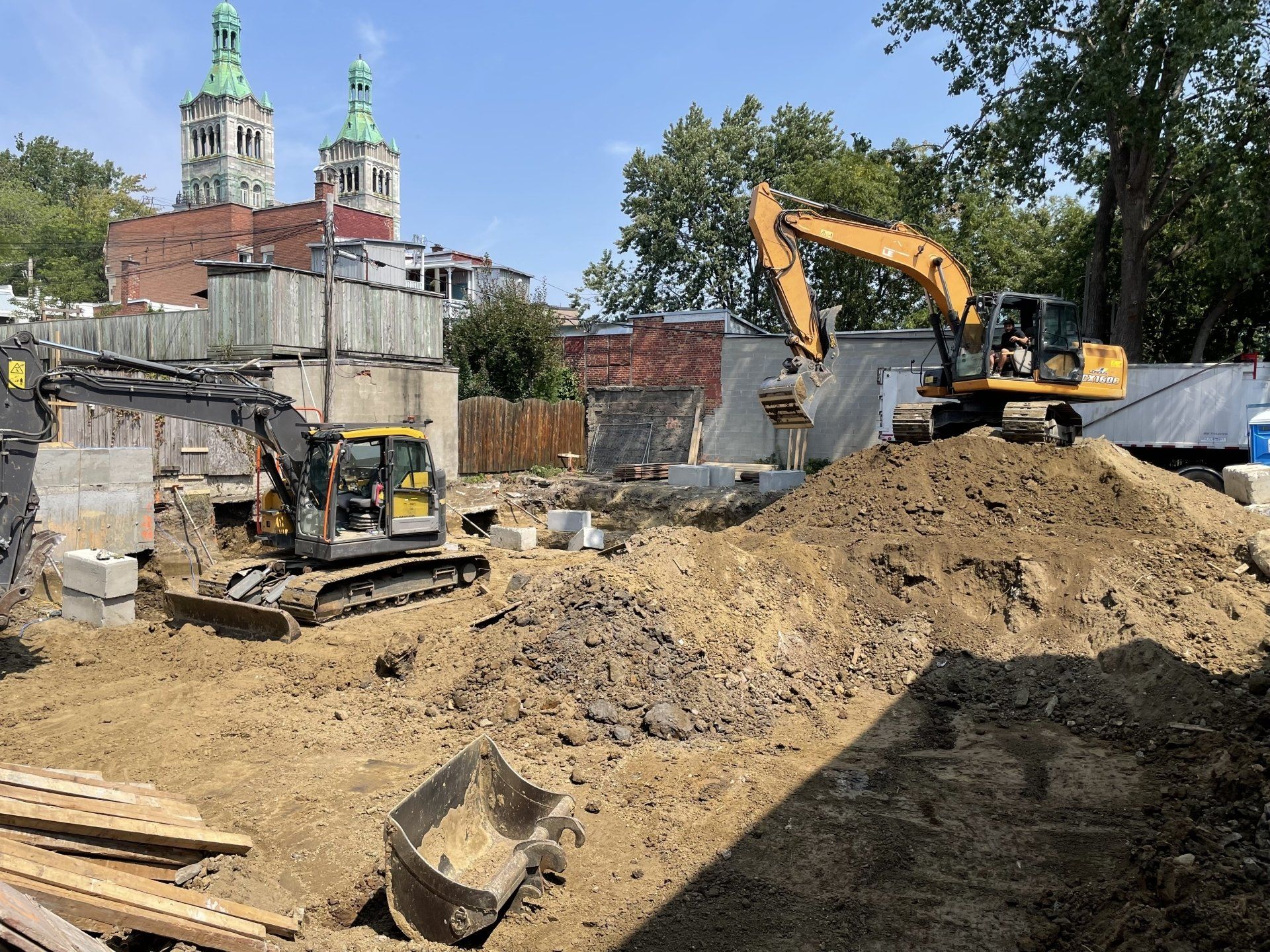 Deux excavatrices sur un chantier, avec une église en arrière-plan. Journée ensoleillée.