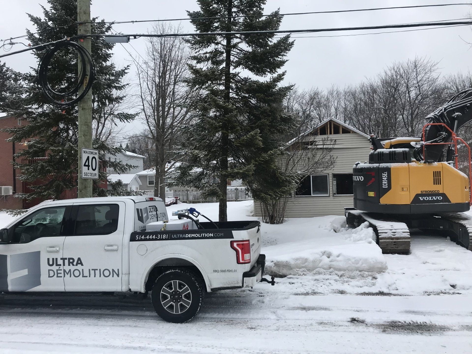Un camion blanc Ultra Demolition et une excavatrice jaune dans une rue enneigée, en train de démolir une maison.