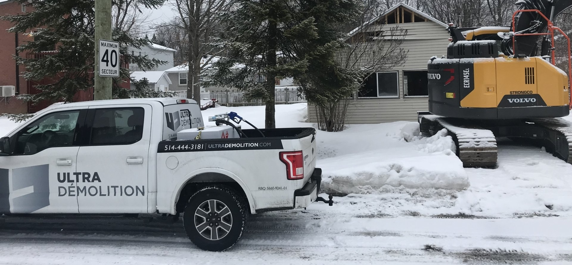Un camion blanc de la société Ultra Demolition et une excavatrice jaune sur un terrain enneigé près d'une maison en cours de démolition.