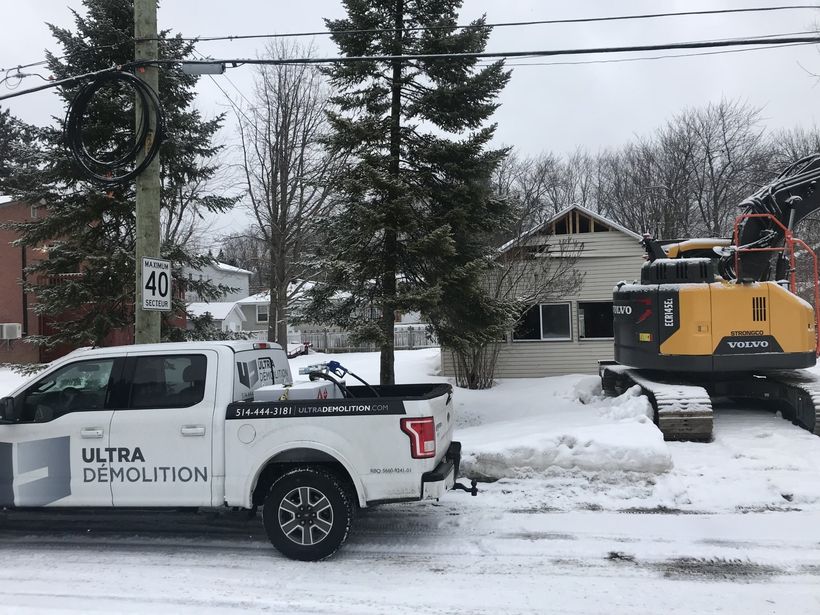 Un camion blanc Ultra Demolition et une excavatrice jaune dans une rue enneigée, en train de démolir une maison.