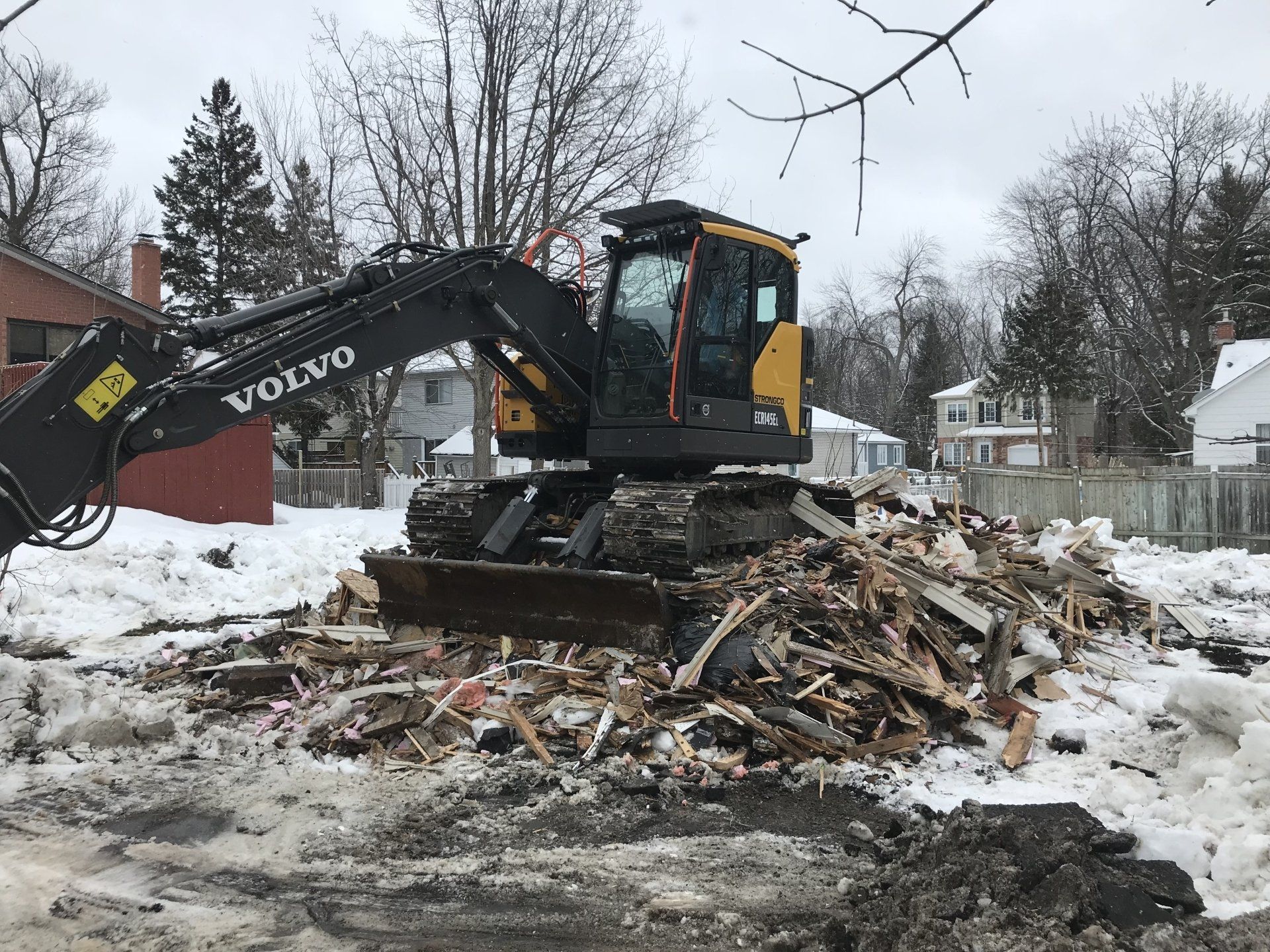 Une pelle mécanique Volvo sur un tas de débris, probablement lors de travaux de démolition dans un quartier résidentiel enneigé.