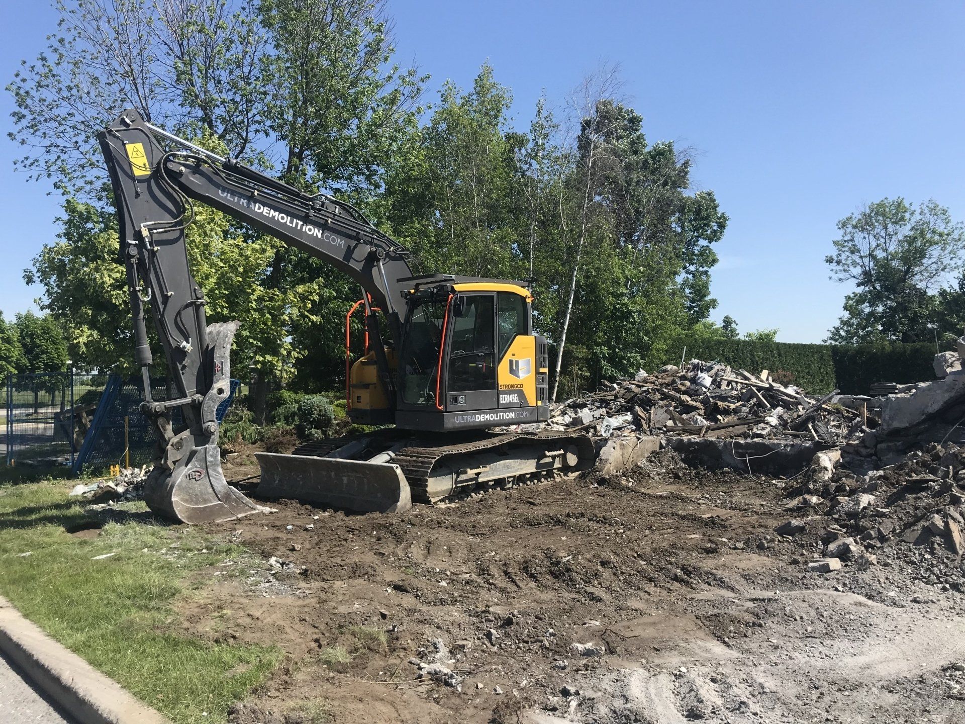 Une excavatrice démolit du béton et des débris sur un chantier. Ciel bleu et arbres en arrière-plan.