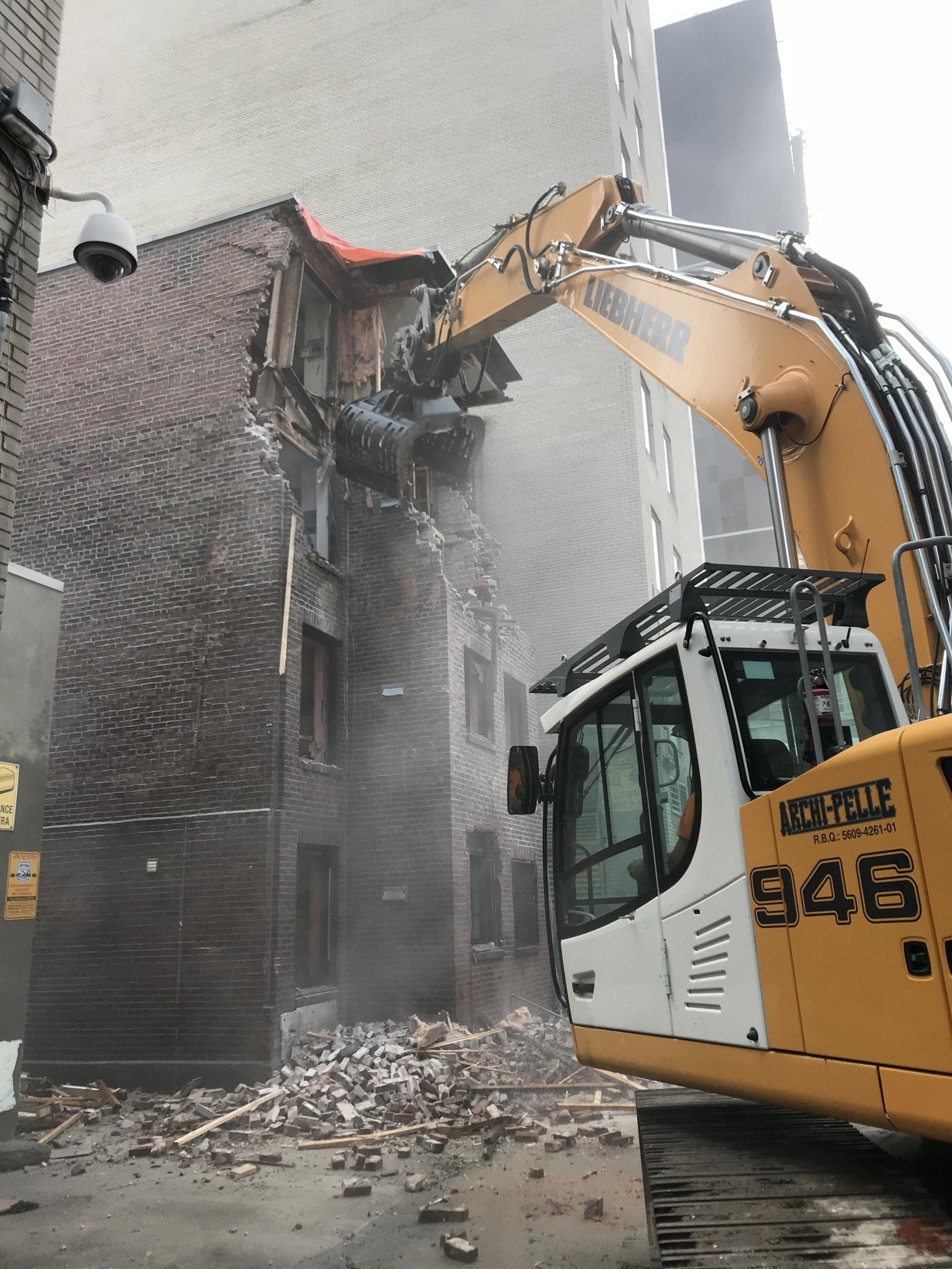 Une pelleteuse démolit un bâtiment en briques ; des gravats jonchent le sol, temps couvert.